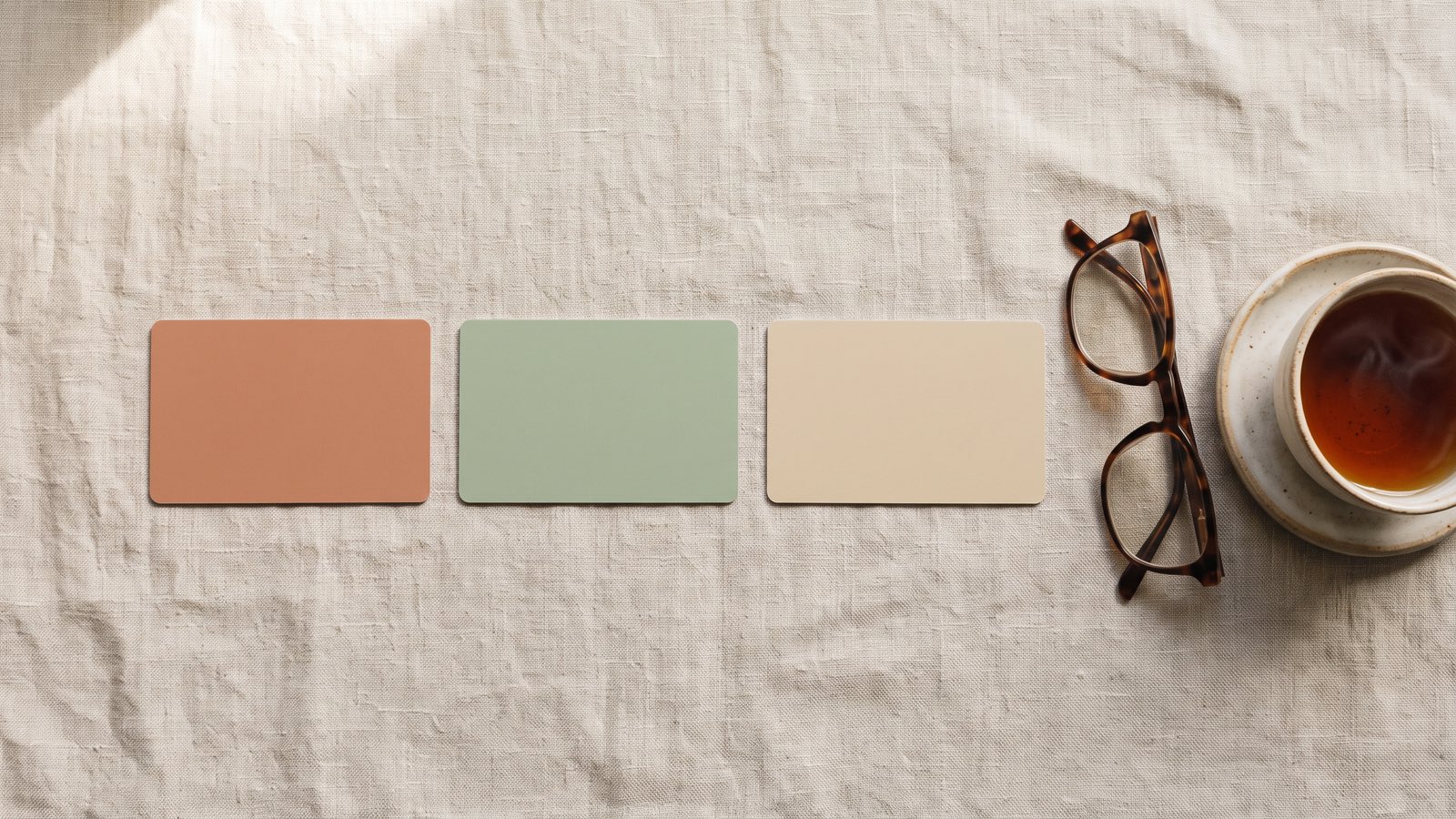 Three blank cards in clay, sage and sand lie apart on a linen cloth beside reading glasses and a cup of tea