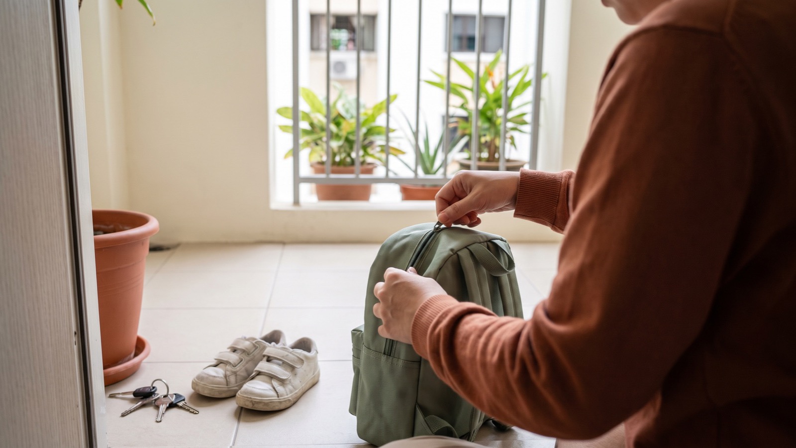 A parent zips a small backpack by the front door of an HDB flat, a pair of white velcro shoes and a set of keys waiting on the floor.